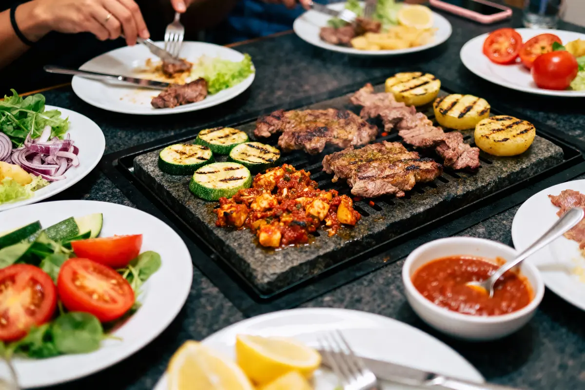 Assiette de grillades, légumes et sauce sur table partagée. Mains avec fourchettes, ambiance conviviale.