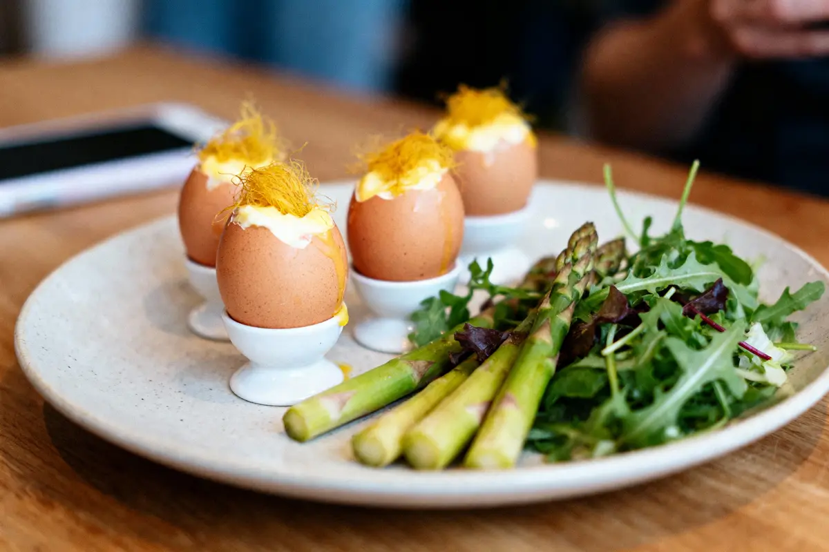Quatre œufs à la coque, salade mixte et asperges dans une assiette blanche sur une table en bois.