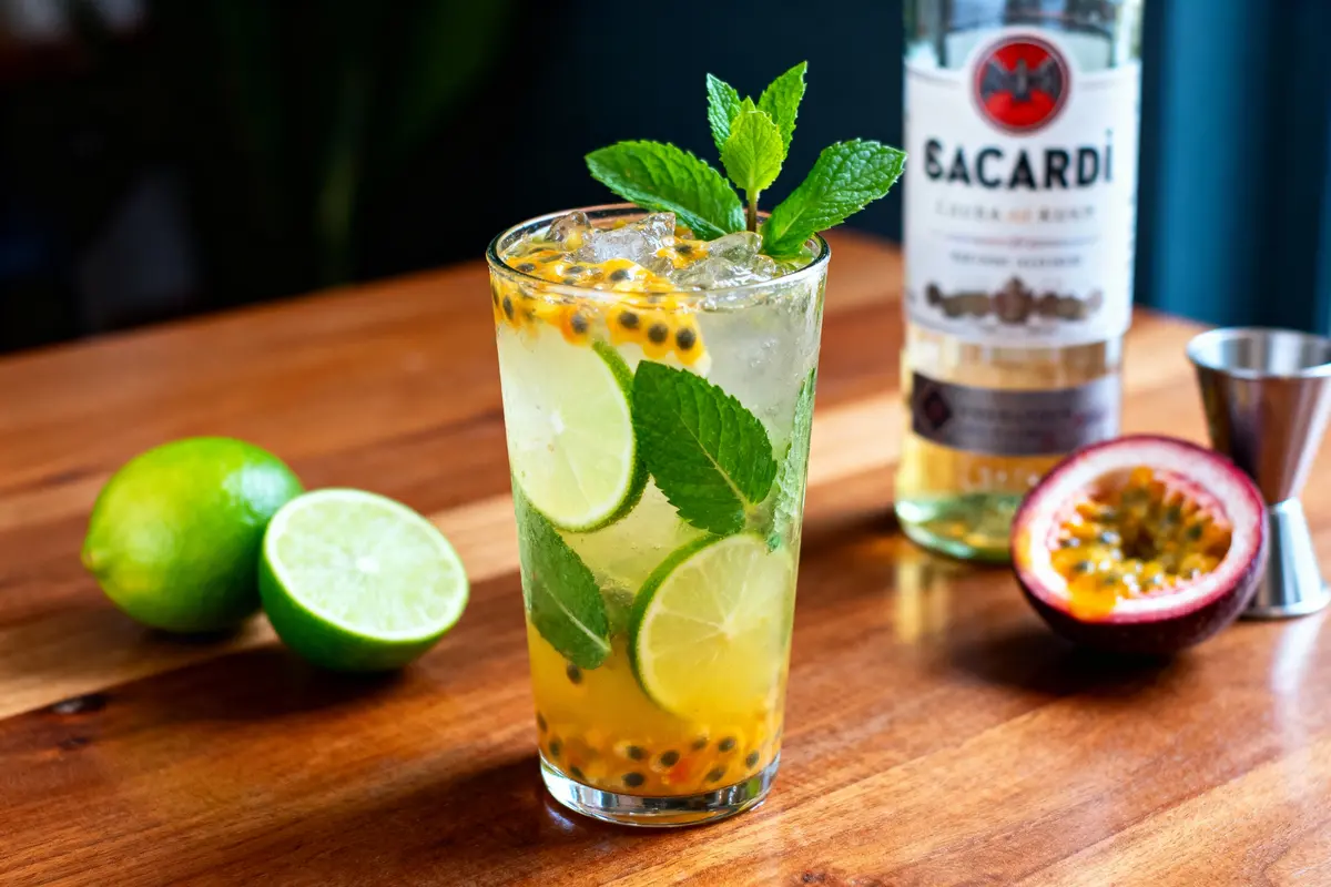 pexels-michal-pech-213601-757318 (1) Verre de cocktail avec menthe, citron vert et fruits de la passion sur table en bois, bouteille de rhum derrière.