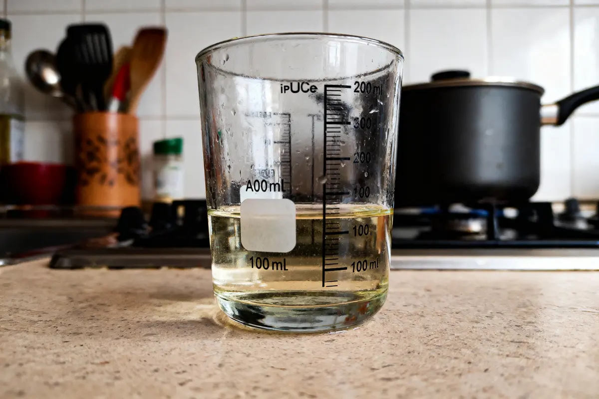 Verre doseur contenant liquide jaune, posé sur comptoir de cuisine; poêle visible en arrière-plan.