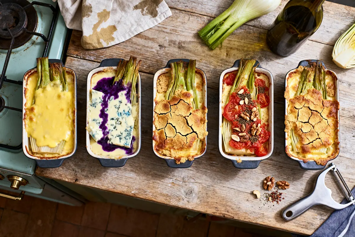 Quatre plats de gratin de fenouil variés sur une table en bois, avec bouteille d'huile et économe.