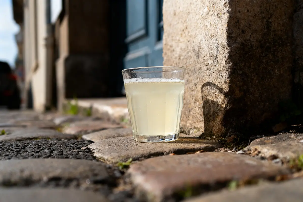 Verre de boisson jaune pâle sur pavés, ombre projetée sur mur ensoleillé.