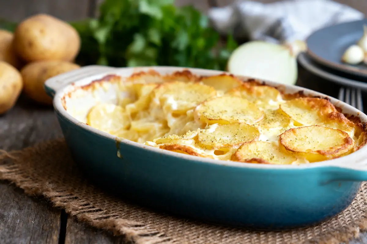 Gratin dauphinois dans un plat bleu sur une table en bois, pommes de terre en arrière-plan.