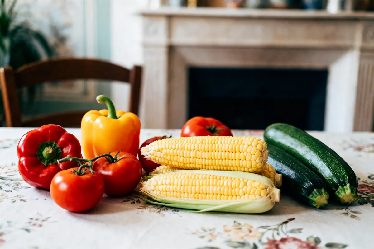 pexels-michal-pech-213601-757318 (1) Légumes variés sur une table : poivrons, tomates, maïs, courgettes, sur nappe fleurie devant cheminée.
