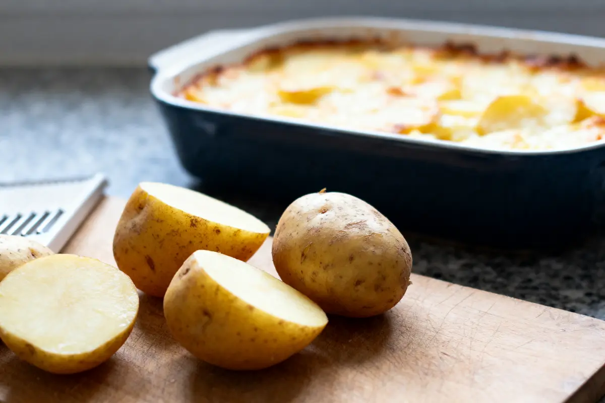 pexels-michal-pech-213601-757318 (1) Pommes de terre coupées sur planche, gratin de pommes de terre au four en arrière-plan.
